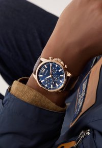 Orologio cronografo color oro al polso, con un quadrante blu, numeri romani, dettagli argentati e un cinturino in pelle marrone.