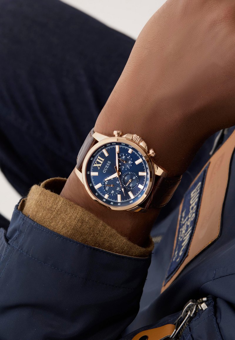 Orologio cronografo color oro al polso, con un quadrante blu, numeri romani, dettagli argentati e un cinturino in pelle marrone.