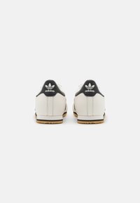 Baskets en cuir blanc avec un accent noir au talon, dotées d'une semelle en caoutchouc et du logo emblématique à trois bandes à l'arrière.