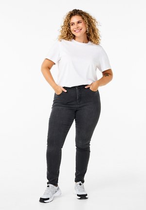 Weißes T-Shirt mit kurzen Ärmeln, kombiniert mit schwarzen Skinny-Jeans; das Model steht mit den Händen in den Taschen und trägt graue Sneakers. Glatte Stoffstruktur.