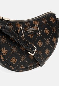 Čierna polkruhová taška Guess s hnedým vzorom loga, zlatým zipsom, zlatou doskou s nápisom "GUESS EST 1981" a nastaviteľným popruhom s prackami.