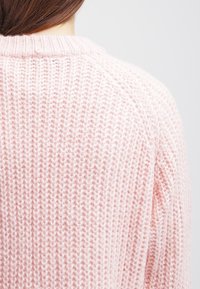 Pull en tricot rose clair avec un motif texturé, présentant un col côtelé et un bouton à l'arrière.