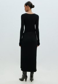 Schwarzes geripptes Strickkleid mit langen Ärmeln, das eine taillierte Silhouette und einen Seitenschlitz aufweist. Getragen mit scharfen schwarzen Stiefeln.