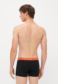 Schwarze Baumwoll-Boxershorts mit orangefarbener Taille, die das Logo "TOMMY HILFIGER" zeigt, von hinten auf einem neutralen Hintergrund dargestellt.