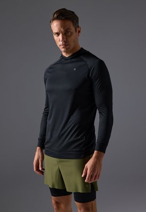 Langemouw zwart sporttop met capuchon, textuurstof en logodetails, gecombineerd met olijfgroene shorts over zwarte compressieshorts.