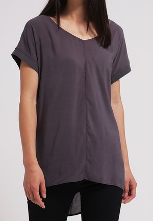 Femme portant un haut ample à manches courtes, col en V gris foncé avec une couture au centre, associé à un pantalon noir.