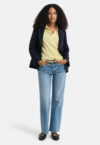 Marineblazer met witte stiksels, gele polo, lichtblauwe jeans en zwarte schoenen met strikken. Model staat met handen in de zakken.