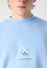 Felpa azzurra con bordo a coste, con il logo "CK" e "Calvin Klein Jeans" in testo bianco a contrasto. Design semplice.