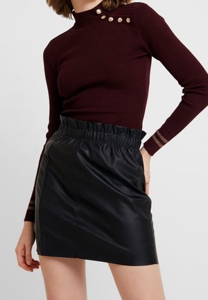 Vrouw die een bordeauxrode geribbelde trui draagt met gouden knopen op de schouder en een zwarte, hooggetailleerde mini-rok van nep leer met een geplooide tailleband.