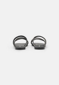 Des sandales plates noires présentent deux straps courbés avec un éclat métallique. La semelle est plate et minimaliste, conçue pour le confort.