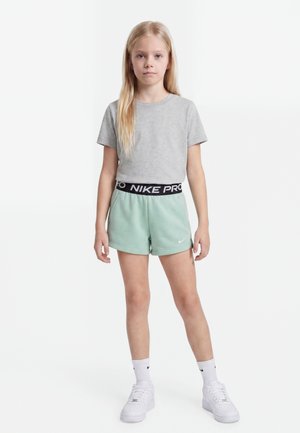 Tüdruk seisab, kandes halli t-särki, piparmündirohelisi Nike lühikesi pükse musta vöökohaga, valgeid Nike sokke ja valgeid tenniseid valgel taustal.