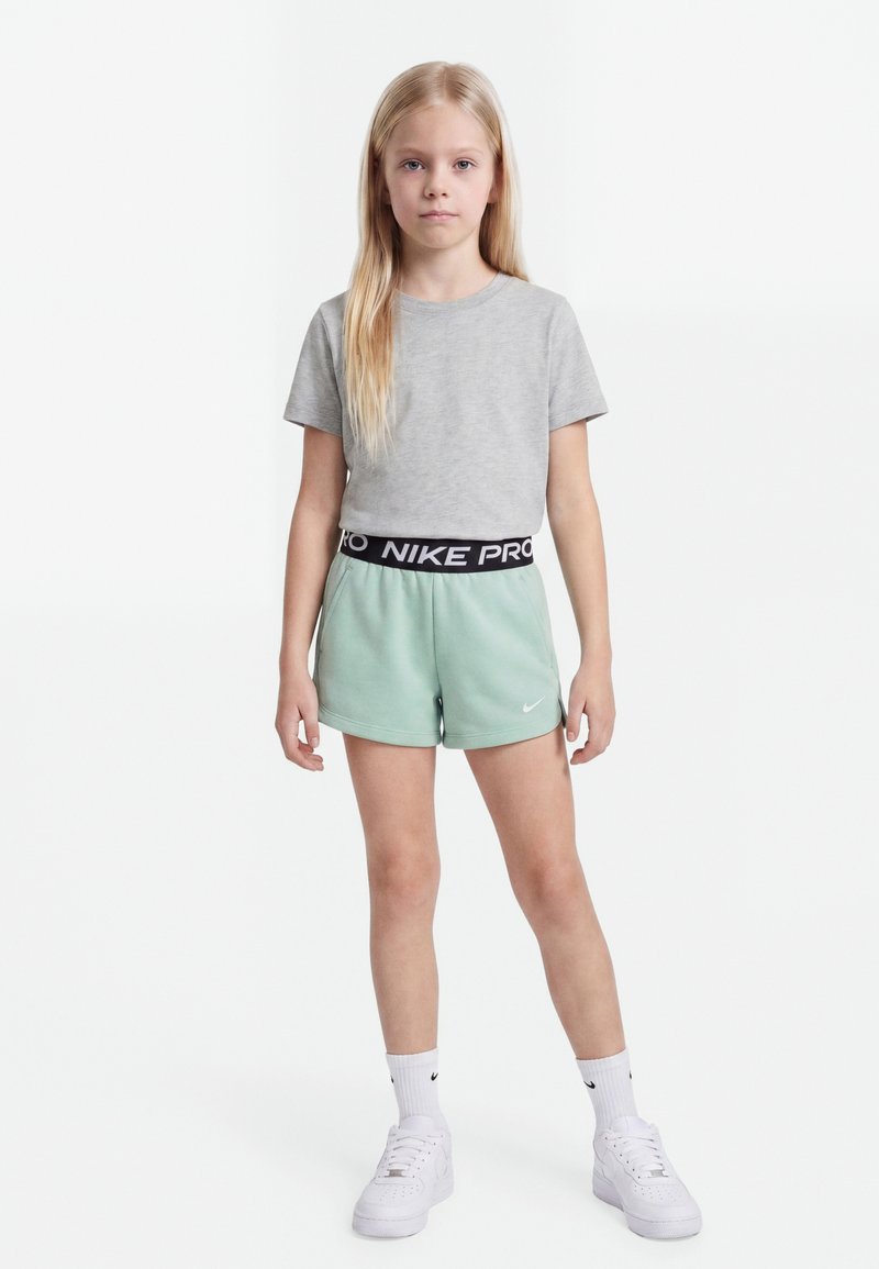 Meisje staand met een grijs t-shirt, mintgroene Nike-shorts met zwarte tailleband, witte Nike-sokken en witte sneakers op een witte achtergrond.