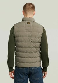 Gilet trapuntato color verde oliva con colletto alto e sezioni cucite; abbinato a una maglietta a maniche lunghe verde scuro e jeans in denim.