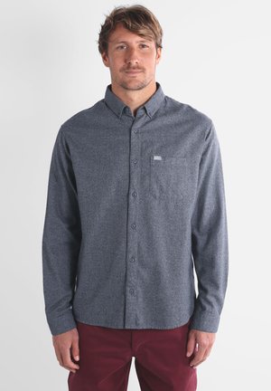 Chemise à manches longues, à boutons, en tissu texturé gris foncé avec une poche poitrine et une petite étiquette. Portée avec un pantalon bordeaux.