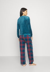 Femme pieds nus portant un haut à manches longues en velours teal et un pantalon de pyjama à carreaux teal, rouge et noir, se tenant contre un fond blanc uni.