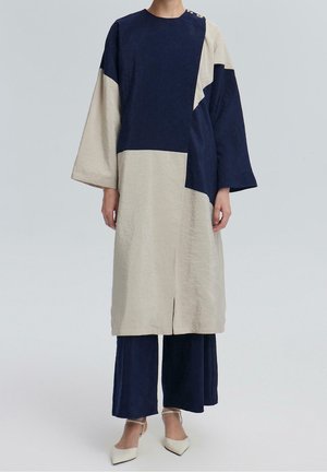 Osoba ubrana w luźną sukienkę i spodnie w blokowe kolory granatu i beżu z długimi rękawami oraz białe szpilki z ostrym noskiem, stojąca na jednolitym tle.