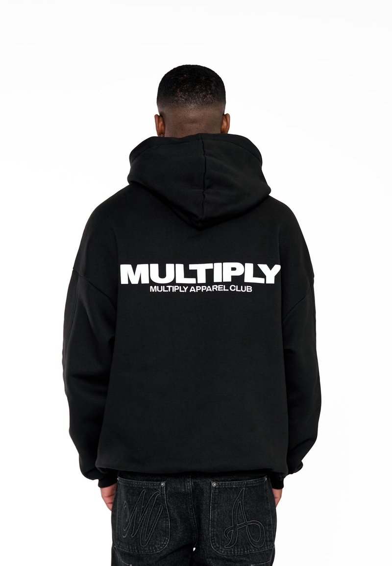 Sweat à capuche noir avec un grand logo blanc "MULTIPLY" au dos. Le matériau semble doux, avec une capuche et une coupe décontractée. Porté avec un jean foncé.