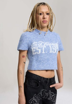 Młoda osoba z blond warkoczami nosząca jasnoniebieski krótki sweter z napisem "Ed Hardy EST. 19" oraz czarne dżinsy z białym haftowanym napisem.