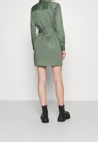 Donna che indossa un vestito corto verde oliva con cintura e stivali neri con lacci alla caviglia, in piedi contro uno sfondo chiaro e semplice.