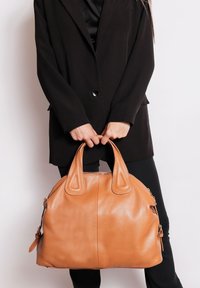 Borsa a mano in cuoio tan, di forma arrotondata, con doppio manico superiore, superficie texture e scomparti con zip. Posizionata su uno sfondo semplice.