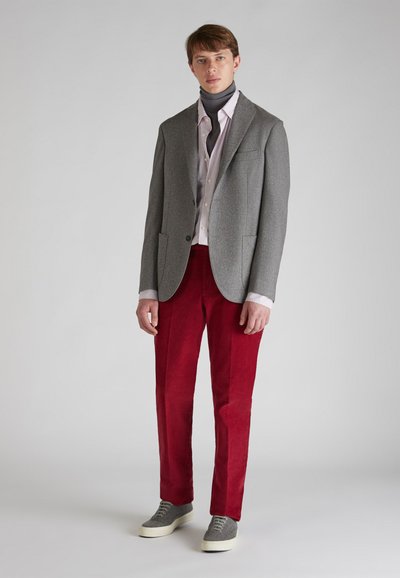 Blazer gris con dos bolsillos frontales, pantalones de pana rojos y zapatillas grises. Con una capa de un suéter de cuello alto oscuro y una camisa de rayas claras.