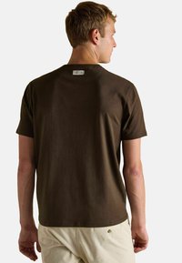 Vīrietis, kurš uzvilcis tumši brūnu īsām piedurknēm t-kreklu un gaiši beigu bikses, redzams no aizmugures uz vienkārša fona.