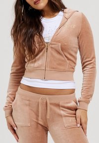 Felpa con zip in velluto tan, decorata con ricami dorati e tasche frontali, abbinata a pantaloni della tuta coordinati con coulisse regolabile.