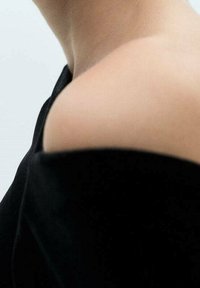 Épaule et cou à découvert portant un vêtement noir à épaule dénudée sur fond uni clair.