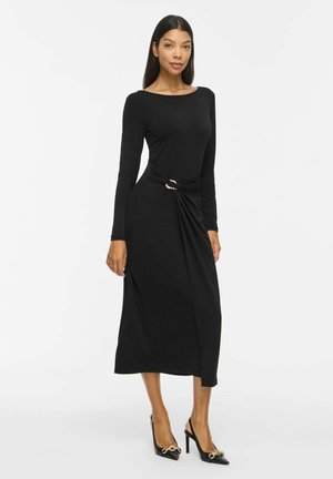 Mannequin portant une robe midi noire à manches longues avec une boucle dorée à la taille et des talons noirs sur un fond clair uni.