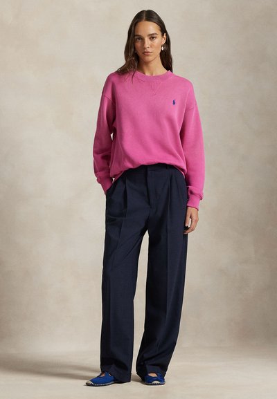 Γυναίκα που στέκεται μπροστά σε ουδέτερο φόντο, φοράει φωτεινό ροζ πουλόβερ, φαρδιά σκούρα ναυτικά παντελόνια και μπλε παπούτσια slip-on, τα χέρια στις τσέπες.