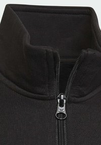 Černá bunda na zip s vysokým límcem, žebrovanou texturou a kovovým zipem s logem. Materiál působí měkce a pružně.