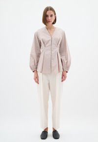Blusa rosa chiaro con scollo a V, dettaglio di bottoni sulla parte frontale, corpino strutturato e maniche a sbuffo; abbinata a pantaloni crema a sigaretta e scarpe nere.