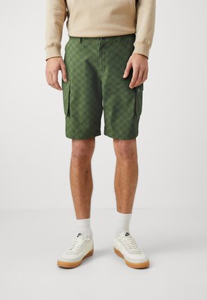 Shorts cargo verzi cu un model texturat, având buzunare laterale mari, închidere cu buton și fermoar, purtați cu adidași albi.