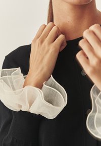 Crna majica s porubom s dugmadi, s prozirnim bijelim ruffles rukavima. Glatka tekstura tkanine, s kontrastom između tamnih i svijetlih elemenata.