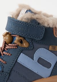 Blå tygsko med bruna snören, fleecekrage och beige detaljer. Har logotyp och texturerad design. Stabil sula för hållbarhet.