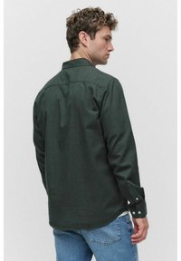 Man met krullend haar, gekleed in een donkergroene lange mouwen shirt en een spijkerbroek, iets naar de zijkant gekeerd tegen een eenvoudige witte achtergrond.