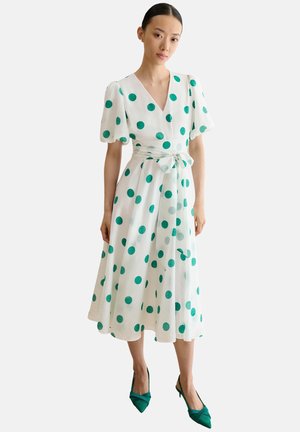 Frau trägt ein weißes Midi-Kleid mit grünen Tupfen, aufgepufften kurzen Ärmeln, einem Wickel-Gürtel in der Taille und grünen spitzen Pumps.