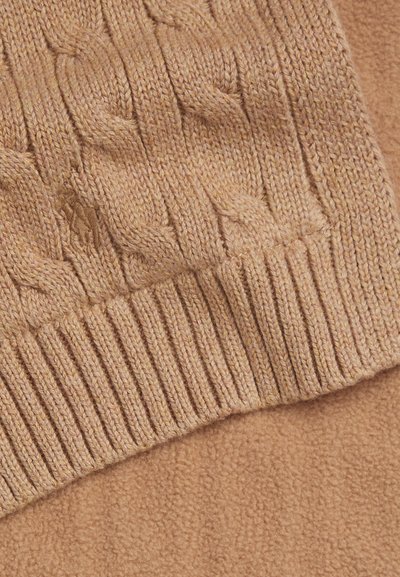 Gros plan sur un poignet de pull tricoté couleur fauve avec un motif torsadé reposant sur une surface en tissu beige doux et texturé.