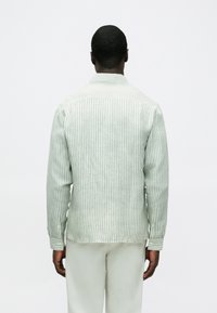 Πουκάμισο με πράσινες και λευκές ρίγες, κατασκευασμένο από ύφασμα με μαλακή υφή. Διαθέτει γιακά και μανσέτες με κουμπιά, με ίσια τελείωμα στο πίσω μέρος.