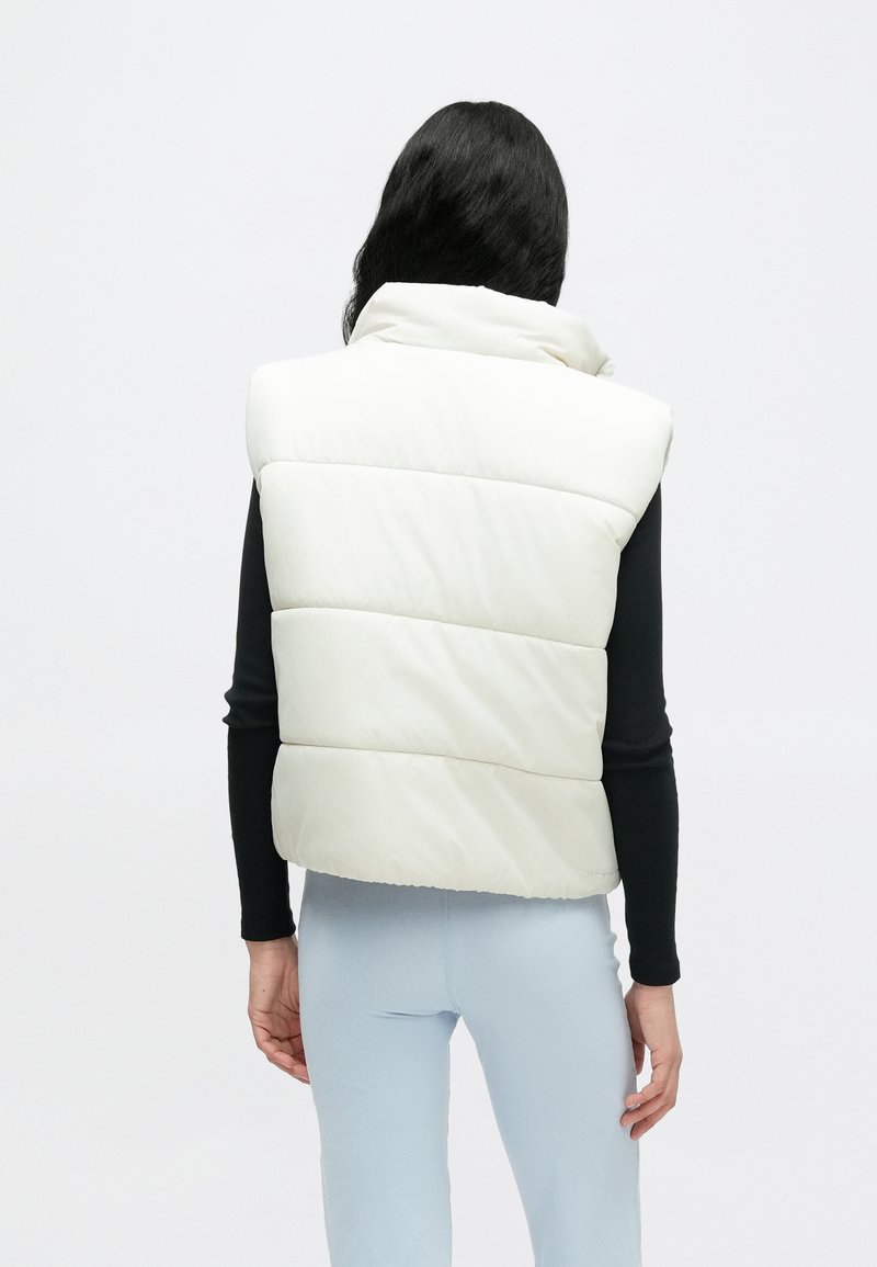 Weiße Steppweste mit hohem Kragen und horizontalen Steppnähten; getragen über einem schwarzen Langarmshirt mit glattem, glänzendem Finish.