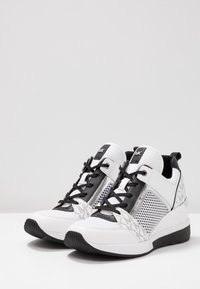 White and black athletic shoes featuring mesh detailing, a smooth leather finish, a cushioned sole, and decorative patterned accents.