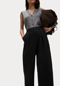Šedý bezrukávový top s knoflíky, spárovaný s černými kalhotami s vysokým pasem a širokými nohavicemi. Modelka drží hnědou semišovou tašku.