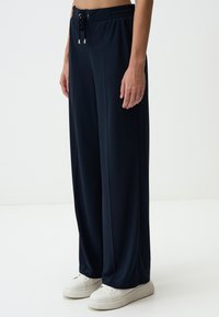 Pantalons larges bleu marine en tissu doux, dotés d'une taille élastique avec cordon de serrage et accents métalliques, associés à des baskets blanches.