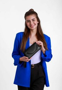 Černá kožená kabelka s vyhnutým klopou, patentním uzávěrem a třemi dekoračními nýty. Modelka drží kabelku a má na sobě modrou sako a černé kalhoty.