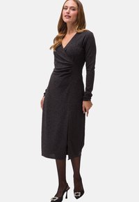 Schwarzes glitzerndes Wickelkleid mit langen Ärmeln, knielangem Saum, seitlichem Rüschen und tiefem V-Ausschnitt, kombiniert mit schwarzen High-Heel-Schuhen.