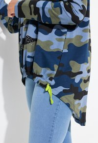 Felpa con stampa camouflage in blu, verde e nero, con orlo regolabile e coulisse verde neon. Tessuto morbido, vestibilità comoda.