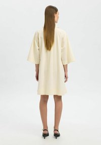 Vestido corte recto color crema con mangas anchas, dobladillo a la altura de la rodilla y textura suave. Se lleva con sandalias de tacón negro.