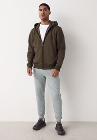 Hanorac cu fermoar verde măsliniu, cu buzunare laterale, combinat cu pantaloni de trening bleu deschis, conici, cu cusături vizibile și tivuri elastice. Tenisi negri completează ținuta.