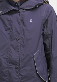 Navy blue waterproof jacket with a hood, featuring a silver logo, button closure, and side pocket outlined in white stitching.