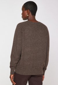 Une personne portant un pull brun tacheté et un pantalon sombre, de dos, avec les cheveux courts et des boucles d'oreilles en or, devant un fond uni.